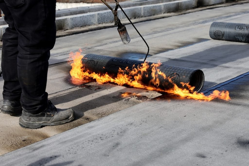 Professional construction workers using a blowtorch to glue modified bitumen roll sheet over a concrete roof.
