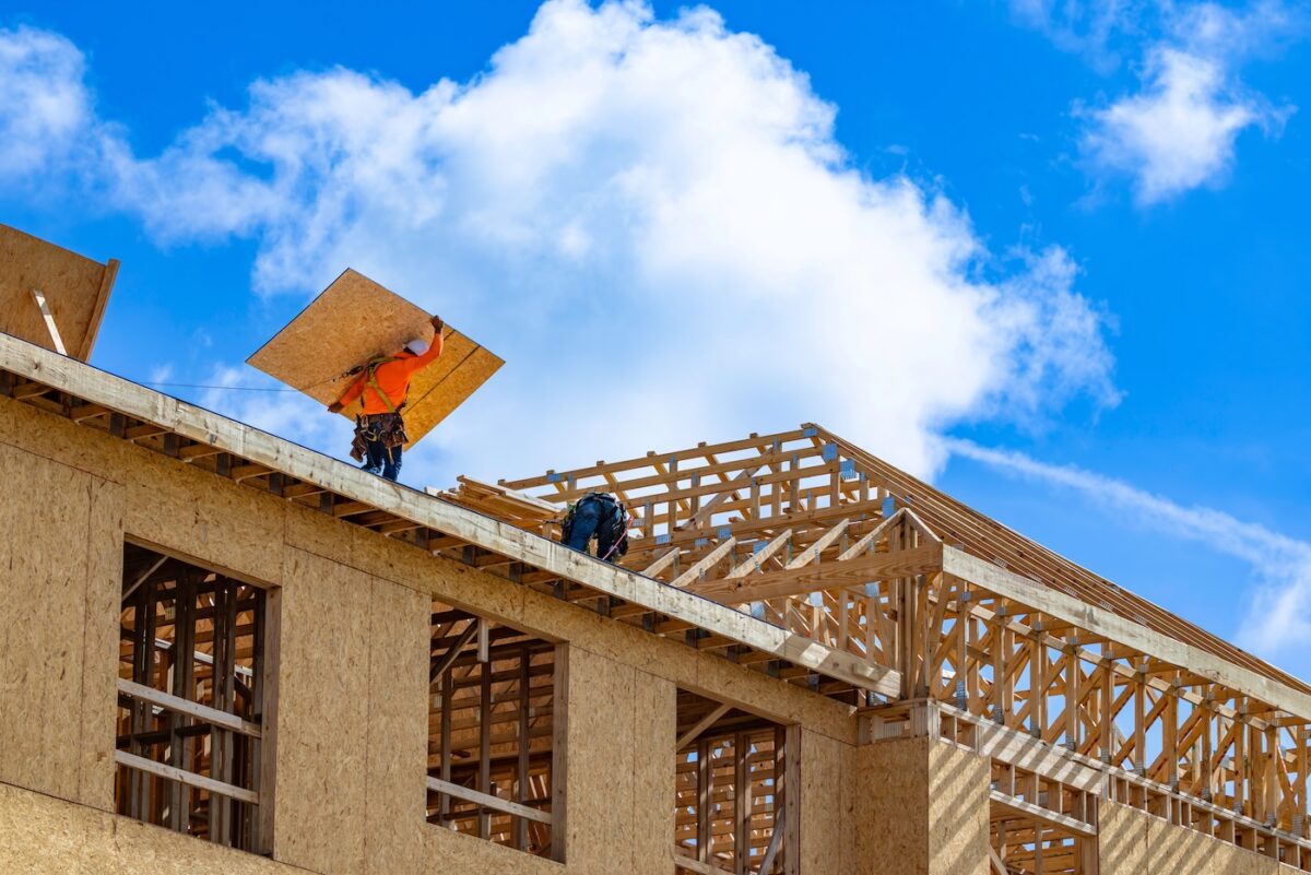 A wide horizontal low-angle view of construction workers wearing safety harnesses while installing plywood sheathing panels over wooden roof trusses