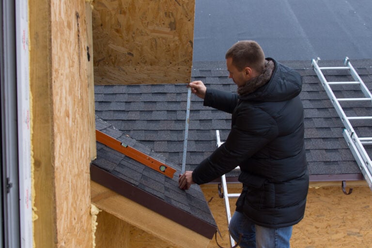 Roofer measuring tiles on a new house with a tape measure as he stands on a ladder while installing the roof